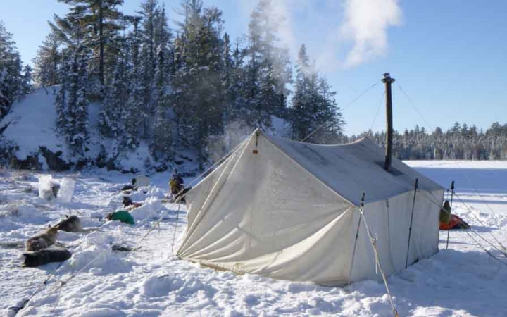 A tent with a small chimney emitting smoke sits on a frozen, snowy lake. Sled dogs rest in the snow nearby. 